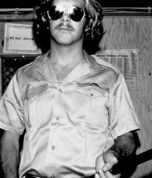 Guard Dave Eshleman, mustached and wearing aviator sunglasses and a khaki uniform, holding a baton.