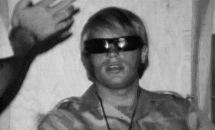 Film still of a guard in mirrored sunglasses seated during the Stanford Prison Experiment.