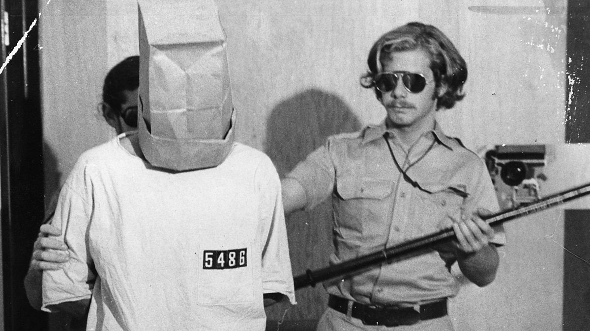 A guard holding a baton escorts a prisoner whose head is covered by a paper bag and whose smock is numbered 5486.