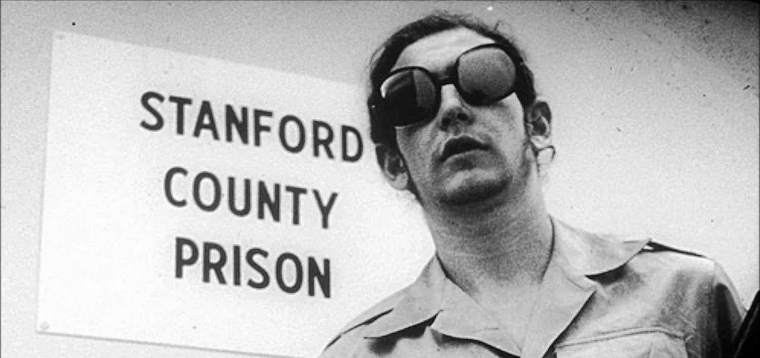A guard in dark sunglasses stands beside a hand-lettered sign reading STANFORD COUNTY PRISON.