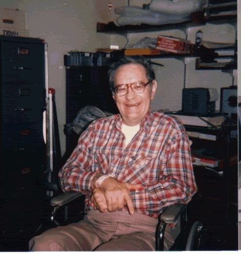A color photograph of an older man, Henry Molaison, smiling while seated in a wheelchair in a cluttered room.