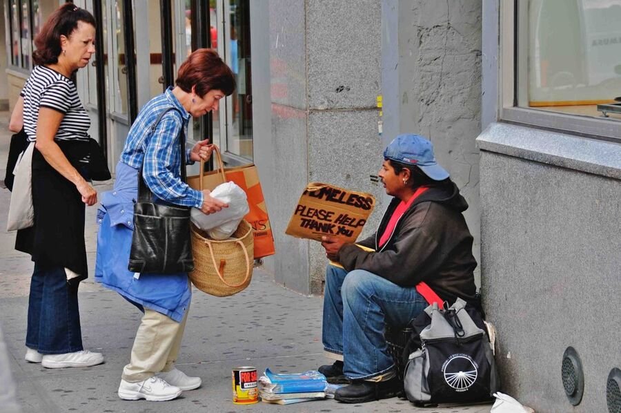 A woman on a city sidewalk bends to hand something to a man sitting against a wall holding a cardboard 'homeless, please help' sign.