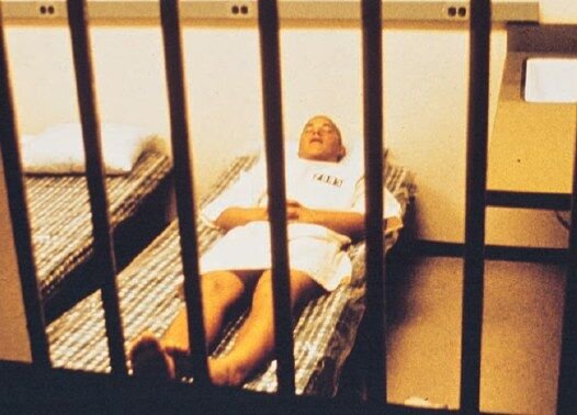 A photo from the Stanford Prison Experiment: a 'prisoner' in a smock lies on a cot inside a barred mock cell.