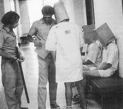 A photo from the Stanford Prison Experiment: 'guards' in uniforms and mirrored sunglasses supervise 'prisoners' who have bags over their heads.