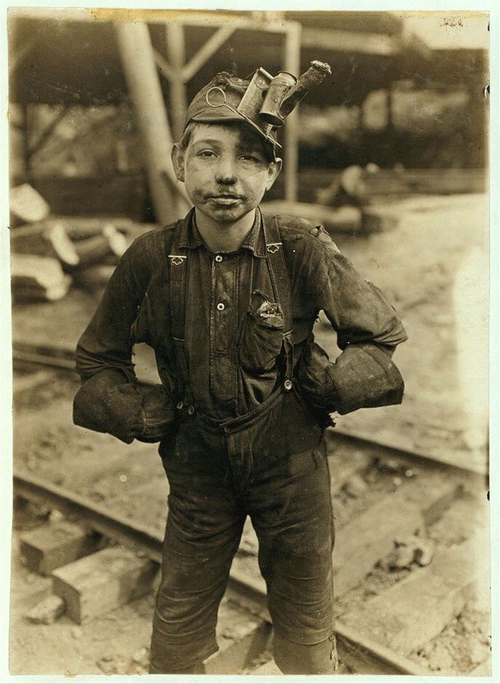 A boy in work clothes standing beside mine tracks with a lamp on his cap.