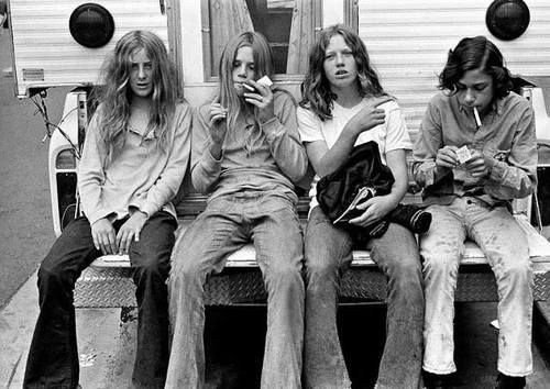 Four teenagers with 1970s hair and clothing sitting together on a porch.