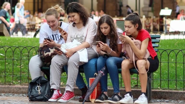 Four teenage girls sitting on a bench, each looking down at a smartphone.